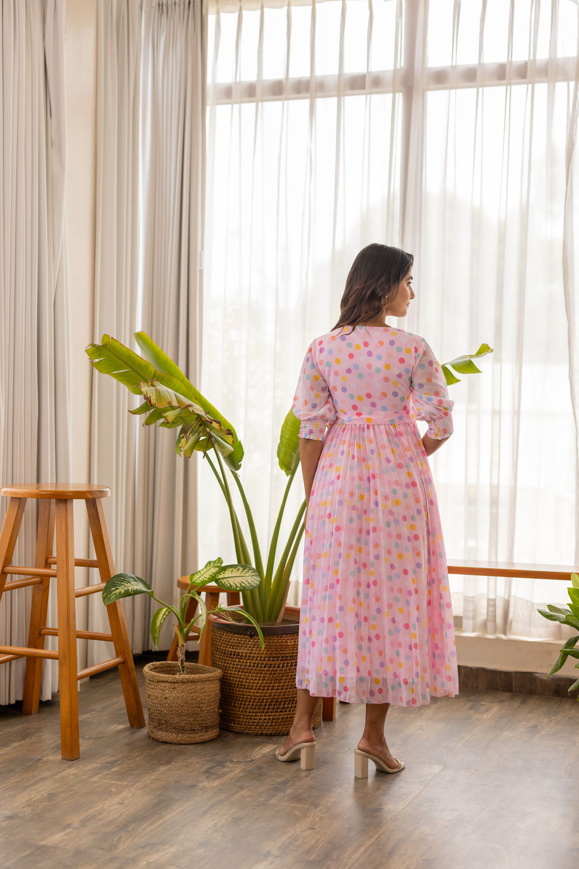 Woman in pink polka pleated maxi dress standing by window.