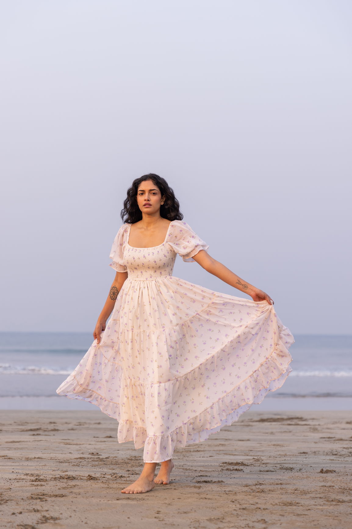 Woman in floral MINIMAL MAXI dress, standing on beach at sunset.