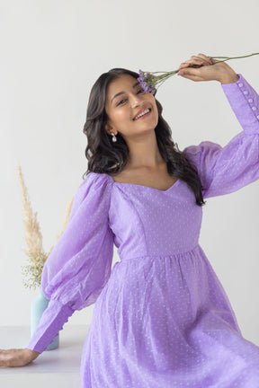 Smiling woman in a lilac princess maxi dress, holding flowers.