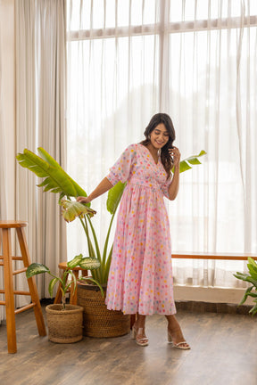 Woman in pink polka pleated maxi dress standing by plants.